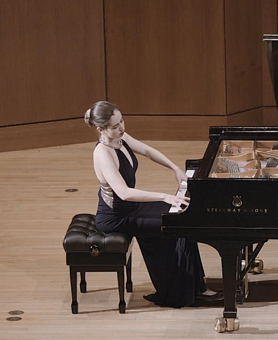 Judith Valerie, wearing a dark blue evening gown, playing a black grand piano on a stage with neutral light brown wood paneling 