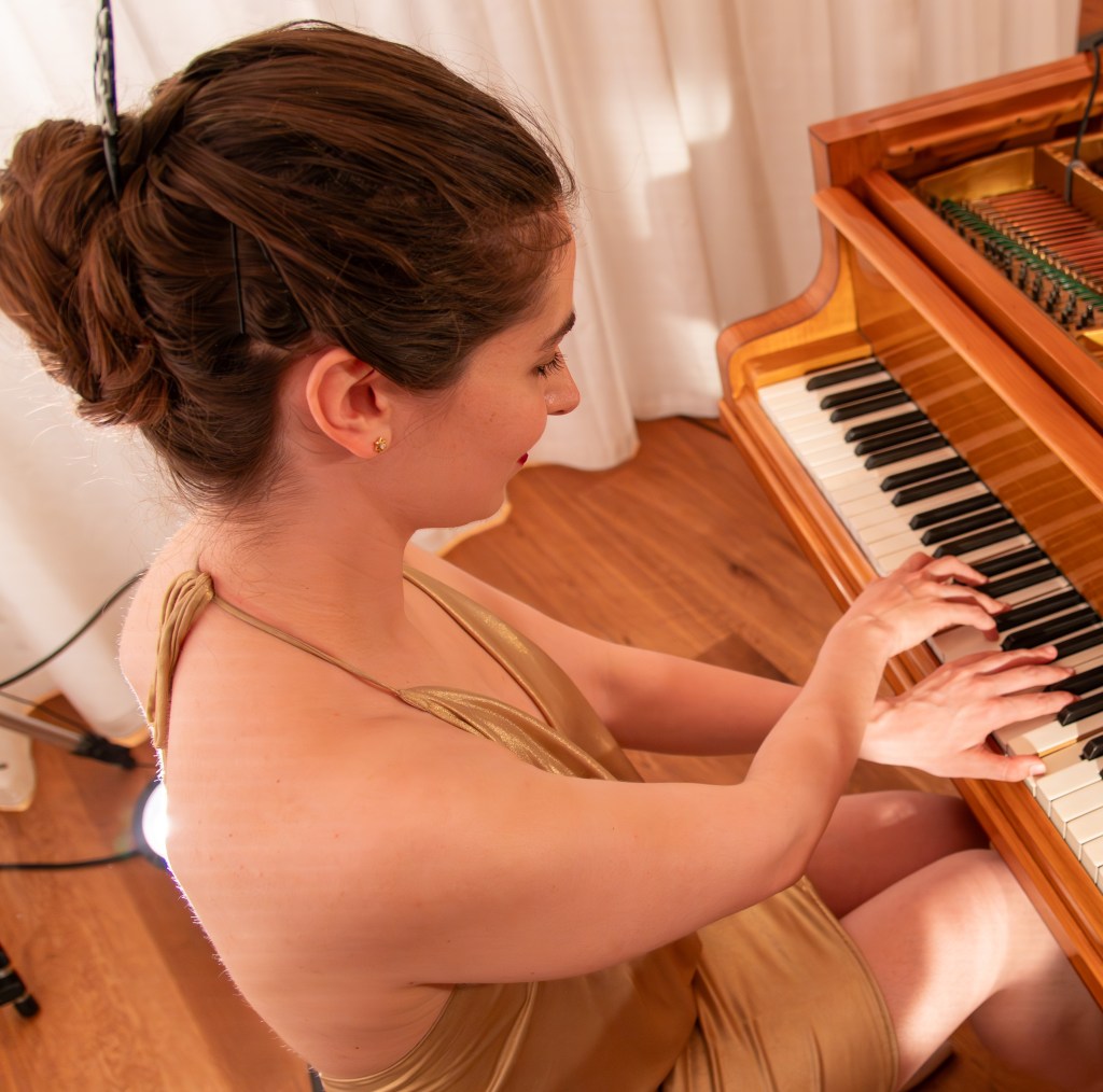Judith Valerie in a golden cocktail dress playing a light brown grand piano, crossing the right hand over the left