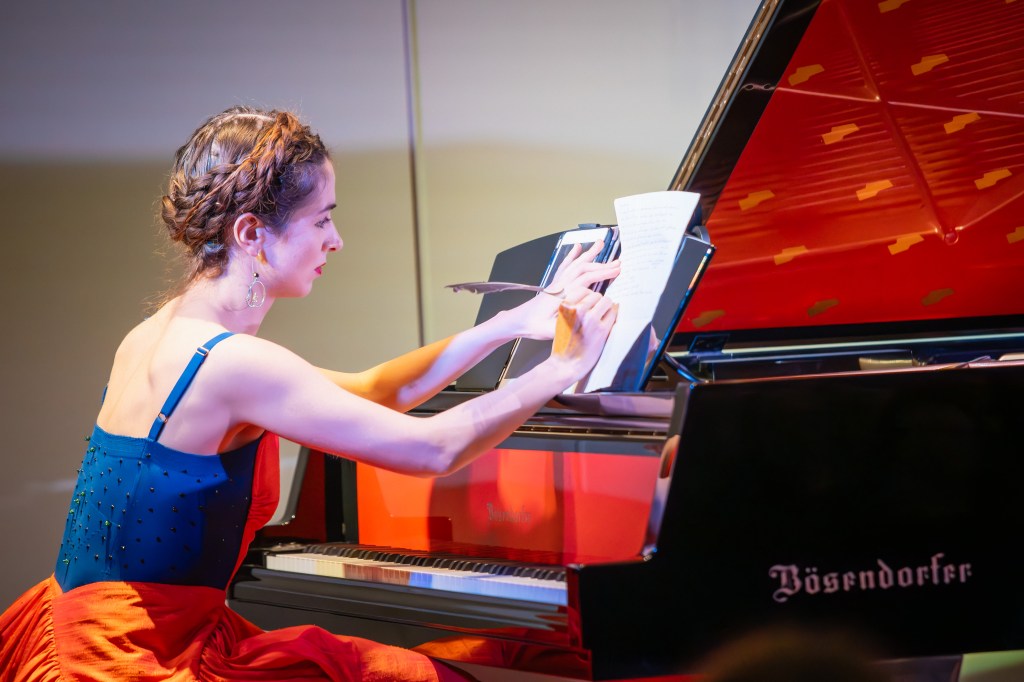 Judith Valerie sitting at a Bösendorfer grand, writing a letter with a feather quill, wearing a red and blue gown by Linda Mafuba; photo © Philipp Gaiko