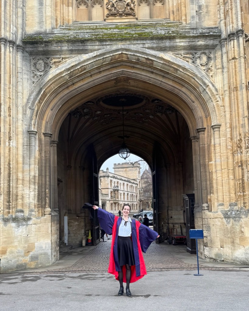 Judith Valerie in red and blue doctoral robes of the University of Oxford in front of the entrance gate of Tom Tower, Christ Church