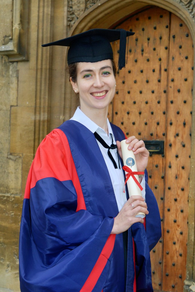 Judith Valerie in an Oxonian red and blue doctoral gown, holding a degree certificate roll